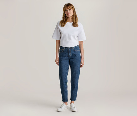 Frau steht, trägt ein weißes Kurzarm-T-Shirt, blaue Jeans und weiße Turnschuhe.