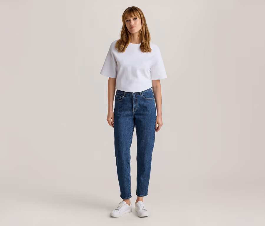 Frau steht, trägt ein weißes Kurzarm-T-Shirt, blaue Jeans und weiße Turnschuhe.