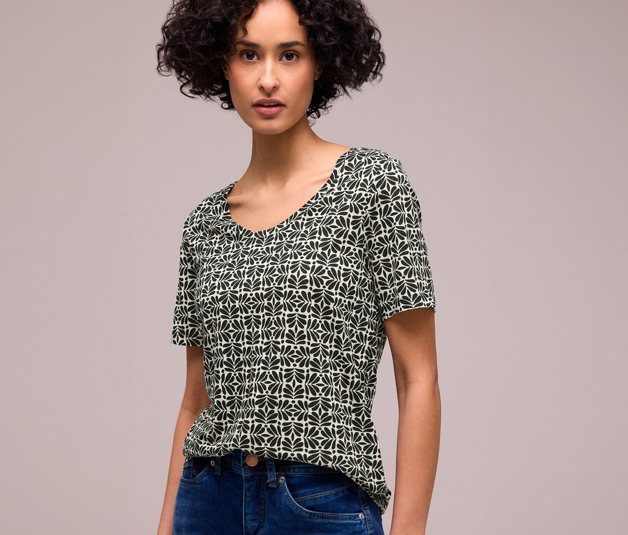 Eine Frau mit dunklem, lockigem Haar trägt ein kurzärmeliges Street One T-Shirt mit schwarzem und hellgrünem Blattmuster und blaue Jeans.