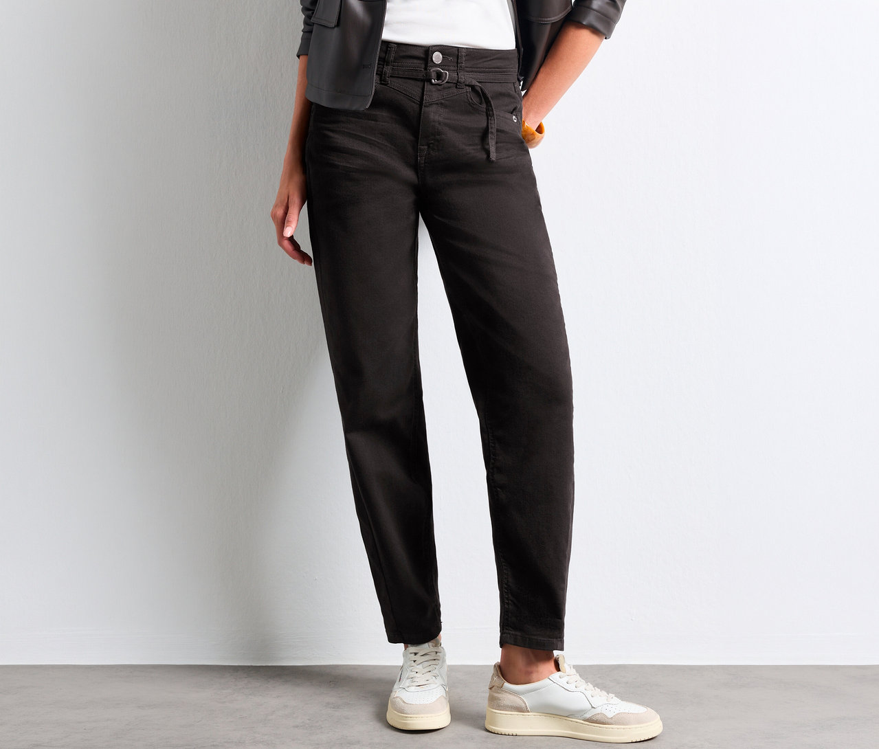 Person trägt schwarze Street One Barrel Jeans mit hohem Bund und Bindedetail, ein weißes Oberteil, eine schwarze Lederjacke und weiße Sneaker.
