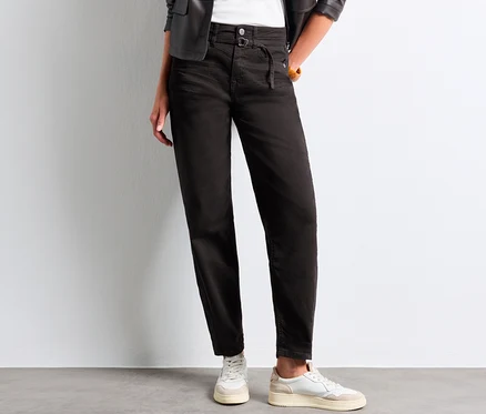 Person trägt schwarze Street One Barrel Jeans mit hohem Bund und Bindedetail, ein weißes Oberteil, eine schwarze Lederjacke und weiße Sneaker.