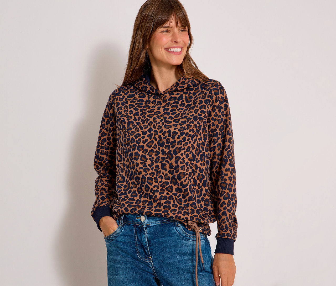 Lachende Frau trägt ein braun-dunkelblaues CECIL Sweatshirt mit Leopardenmuster und Kapuze sowie blaue Jeans, Hände in den Taschen.