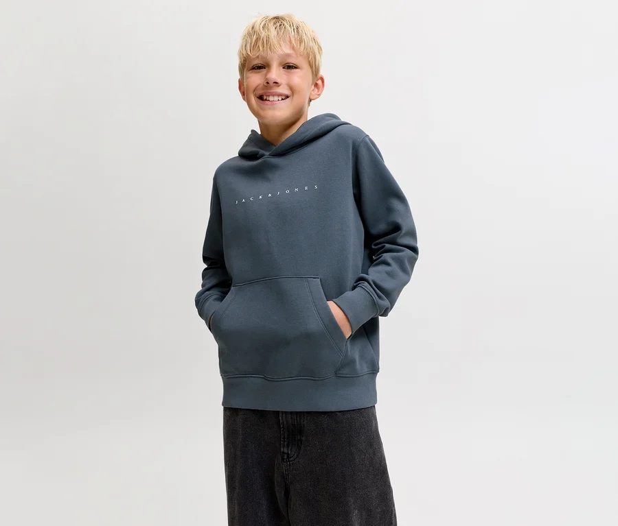 Lächelnder Junge trägt einen dunkelgrauen Jack&Jones Sweat-Hoodie Star und dunkle Hose, Hände in der Kängurutasche.