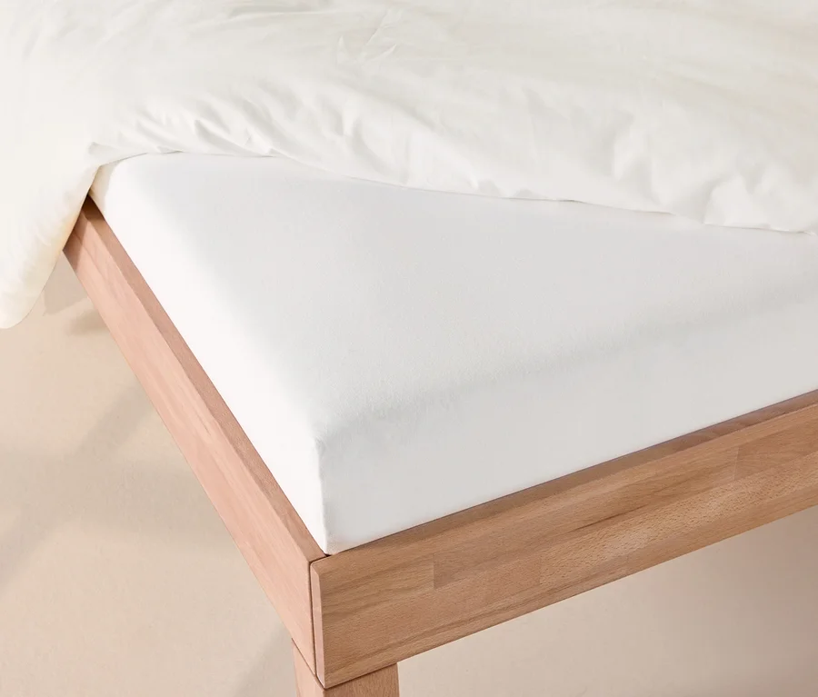 Weißes Jersey-Spannbettlaken auf einem Holzbett, mit einer weißen Bettdecke darüber.
