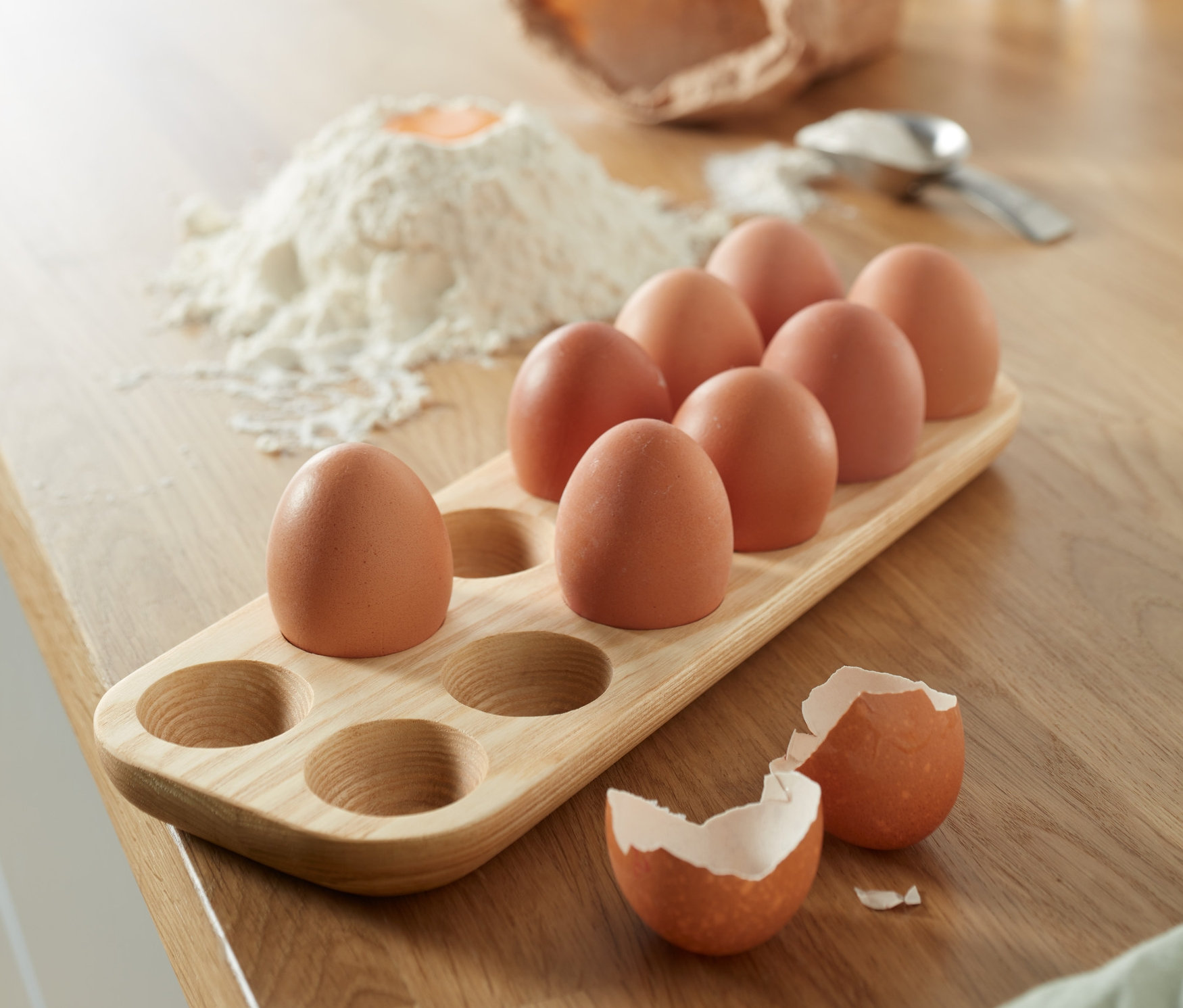 Braune Eier in einem Eierhalter aus Holz, daneben ein Mehlhaufen und eine zerbrochene Eierschale.