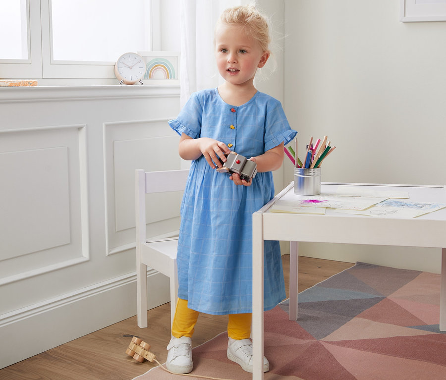 Mädchen hält Spielzeug, trägt ein Webkleid aus Bio-Baumwolle und steht an einem weißen Tisch.