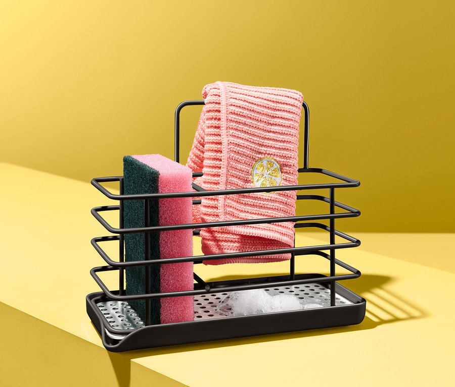 Schwarzer Spülutensilien-Ständer mit Schwamm und Geschirrtuch auf gelbem Hintergrund.