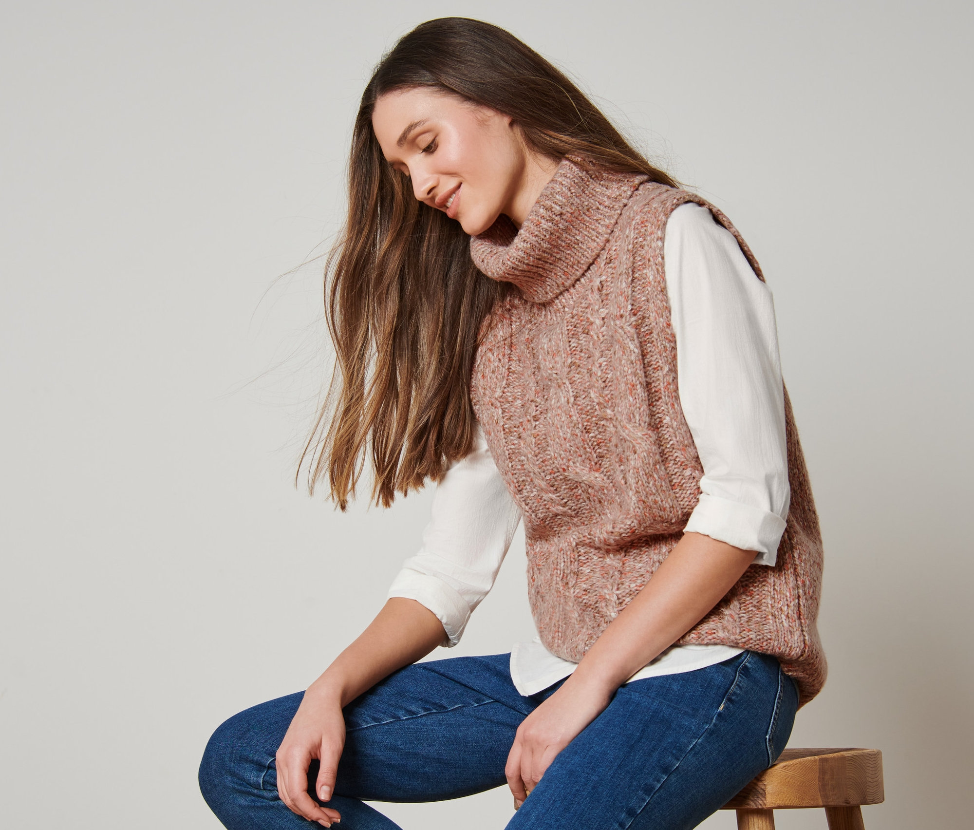 Frau sitzt auf einem Hocker und trägt einen Strickpullunder mit Zopfmuster.