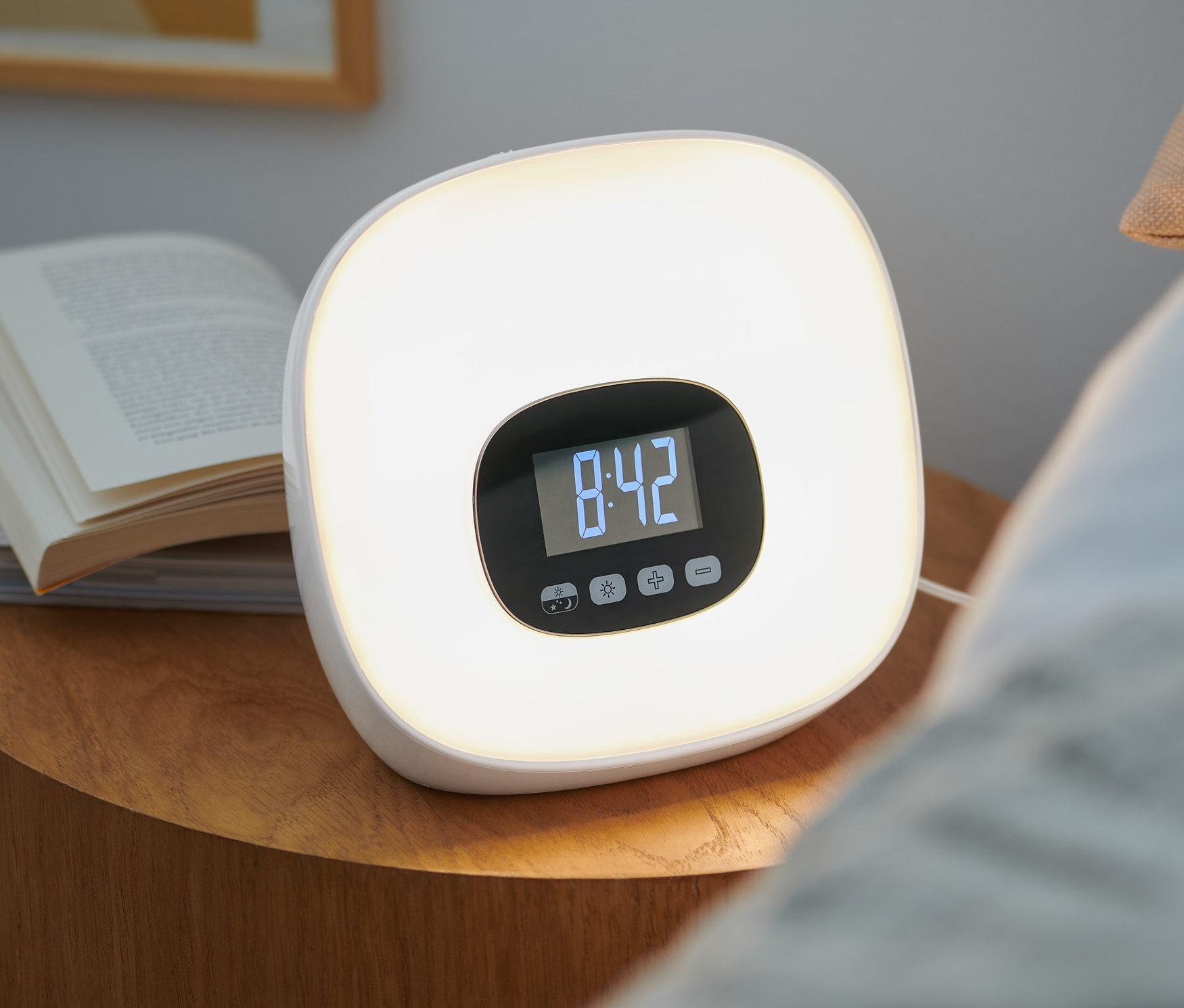 Ein Lichtwecker mit Radio steht neben Büchern auf einem Nachttisch. Auf dem Display des Weckers wird die Uhrzeit 8:42 angezeigt.