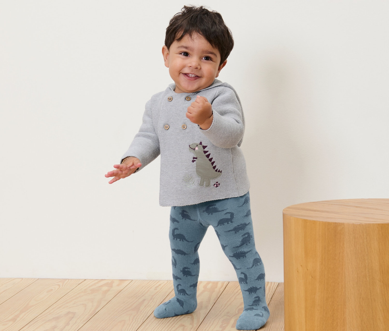 Ein lächelndes Kleinkind steht und trägt eine blaue Strumpfhose mit Dinosaurier-Muster.