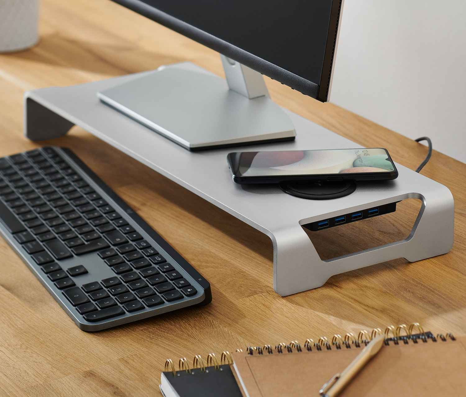 Monitor steht auf Monitorständer mit kabelloser Ladestation und USB-Hub. Auf dem Ständer liegt ein Handy. Daneben steht eine Lampe.