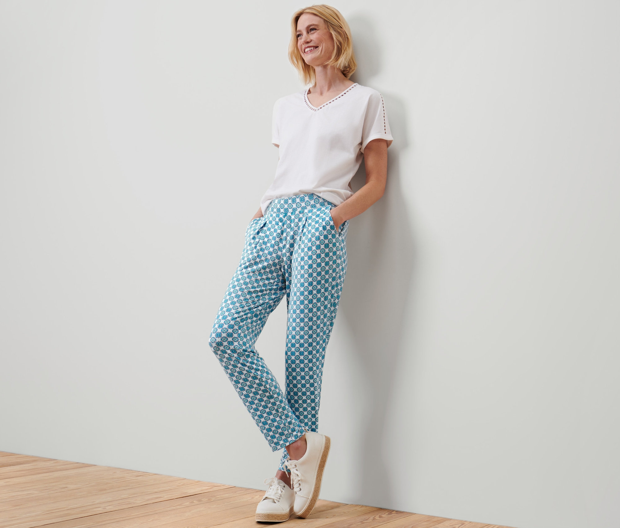 Frau lächelt an eine Wand gelehnt und trägt eine Jerseyhose mit Alloverprint.