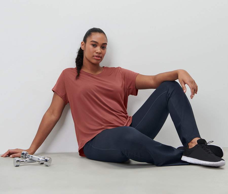 Frau sitzt in terrakottafarbenem Funktionsshirt und Bodyforming-Sporthose neben Hanteln auf dem Boden.