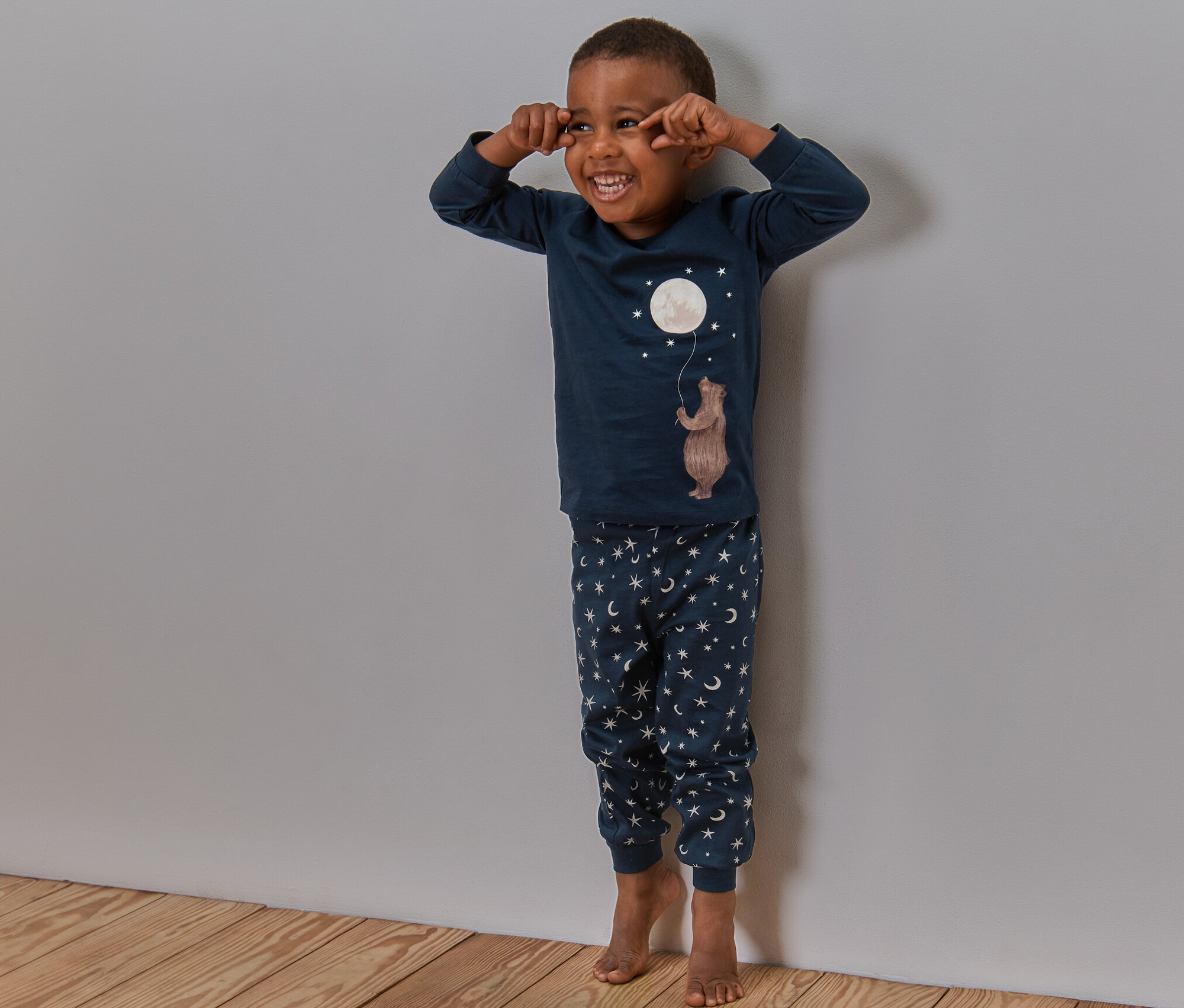 Ein Junge lacht und steht in seinem Kinder-Pyjama »Mond«, glow in the dark an der Wand.