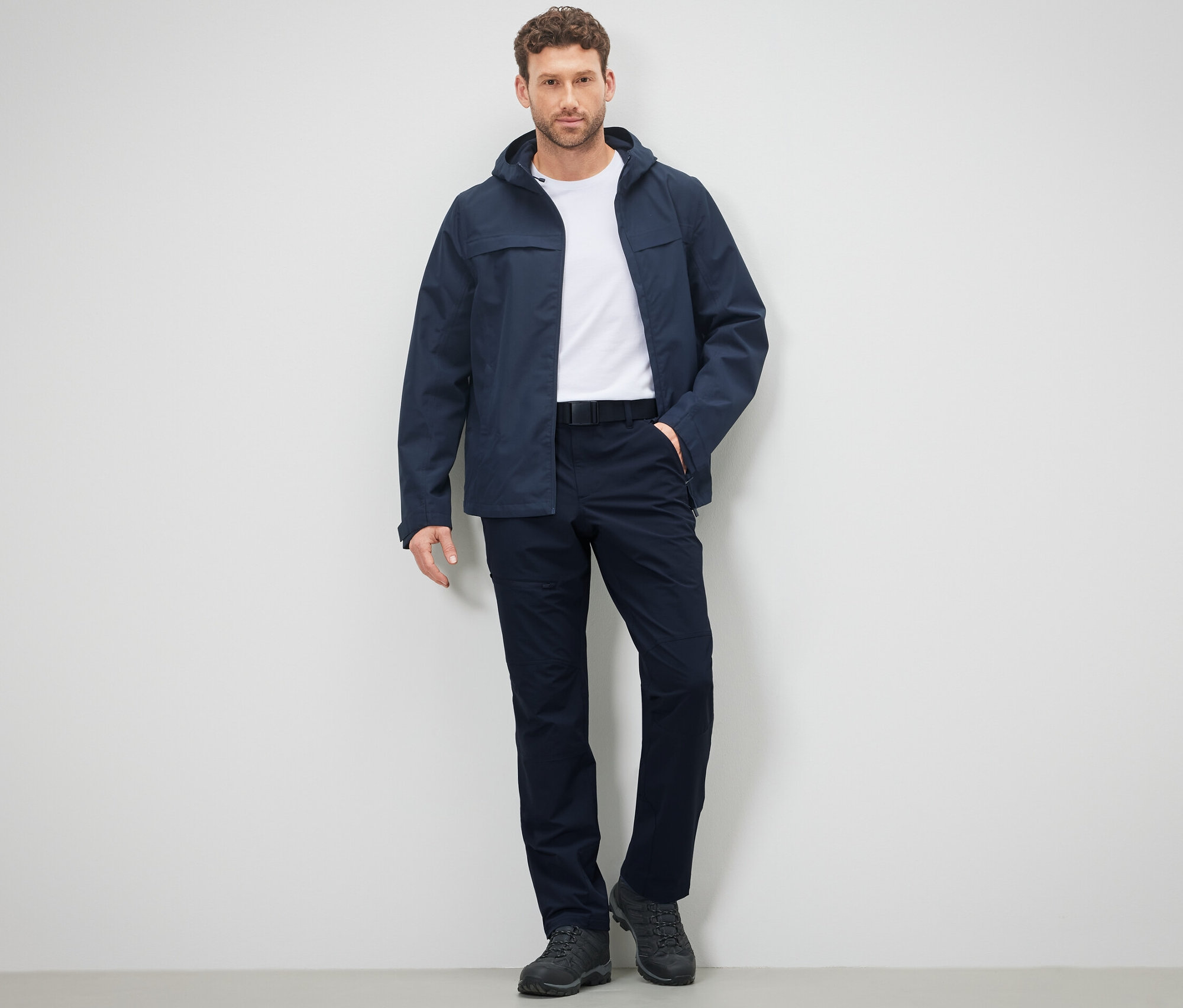 Mann posiert in Outdoorhose und Jacke vor einer Wand.