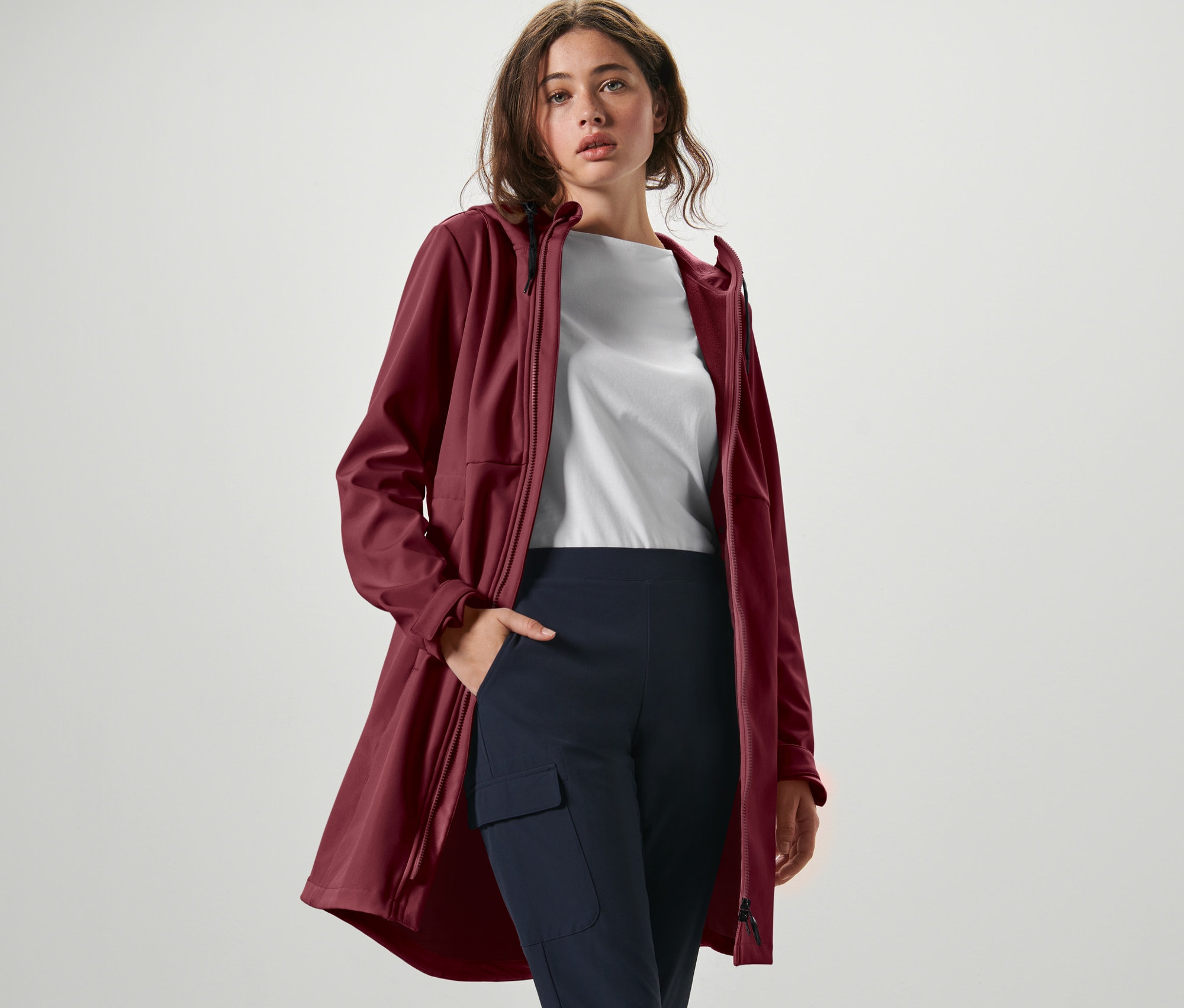 Frau posiert in weinrotem Softshellmantel, weißem T-Shirt und marineblauer Hose.