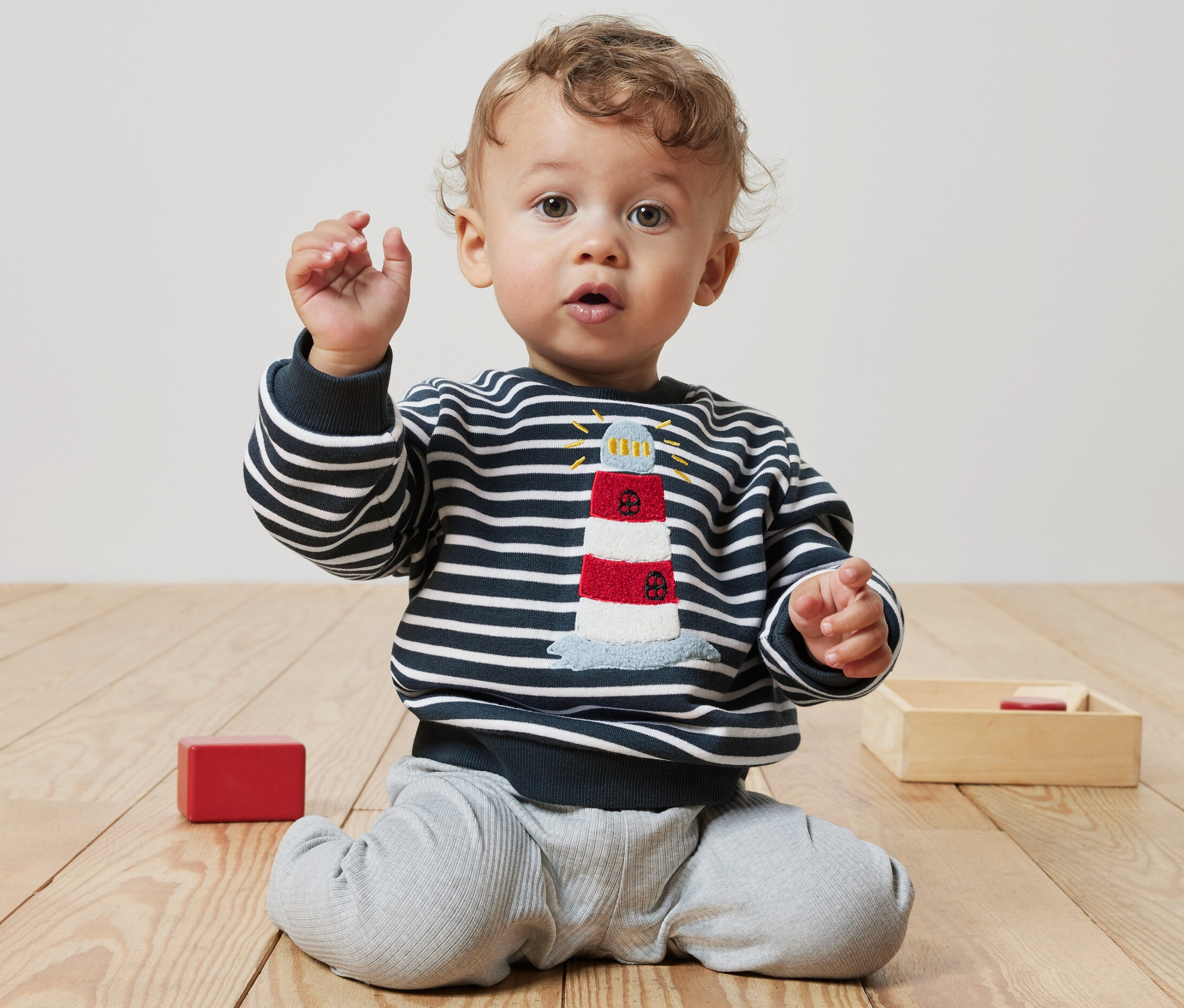 Ein lockiges Baby sitzt auf dem Boden und trägt ein Baby-Sweatshirt mit Leuchtturmmotiv.