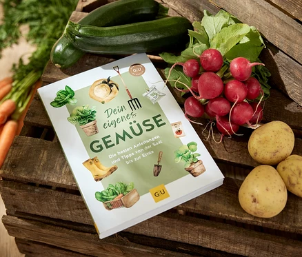 Das Buch »Dein eigenes Gemüse« liegt auf einer Holzkiste, umgeben von Karotten, Zucchini, Radieschen und Kartoffeln.