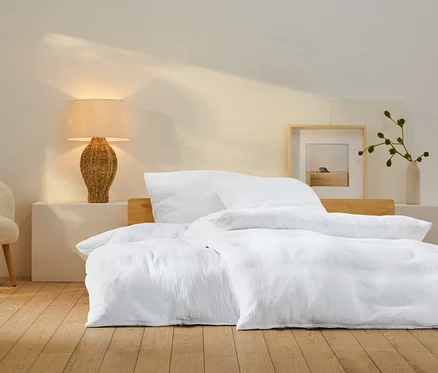 Schlafzimmer mit weißer Bettwäsche auf dem Bett, einer Lampe aus Korbgeflecht und einem beigen Sessel.
