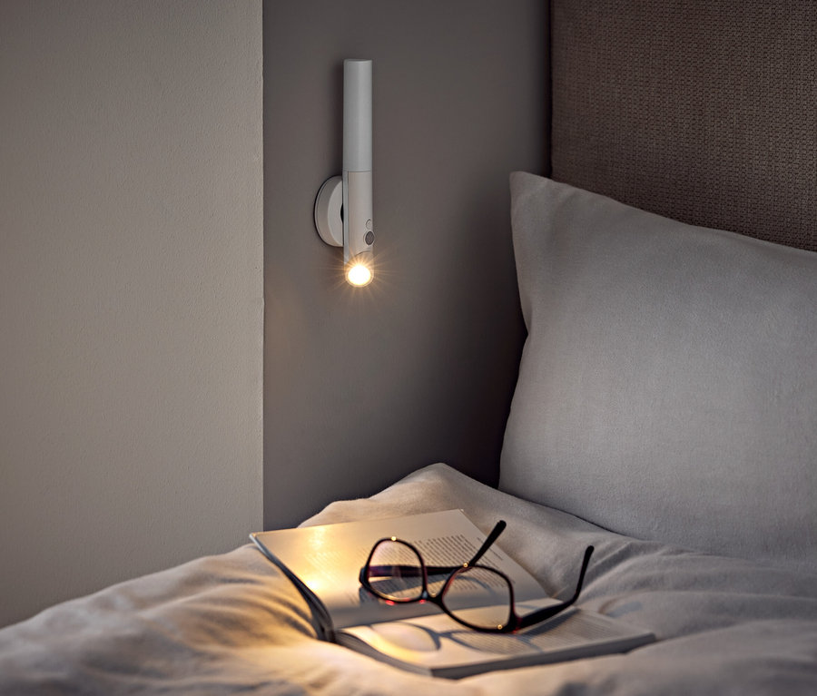Auf einem Bett liegen eine Brille und ein offenes Buch unter einer Wandlampe.