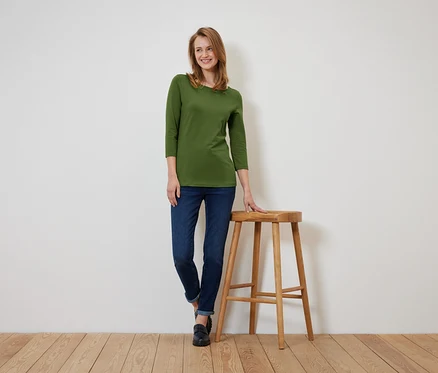 Eine Frau in einem olivgrünen Dreiviertelarm-Shirt und dunkelblauen Jeans steht und lehnt sich an einen Holzhocker.