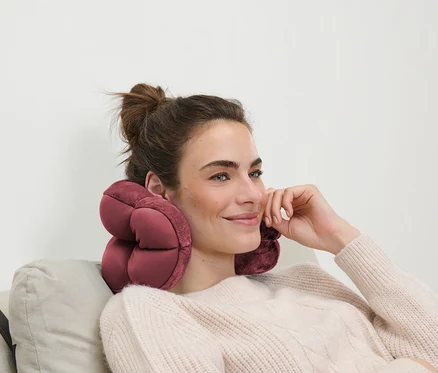 Frau mit braunen Haaren sitzt auf einem Sofa mit einem rosa Reisekissen um den Hals.