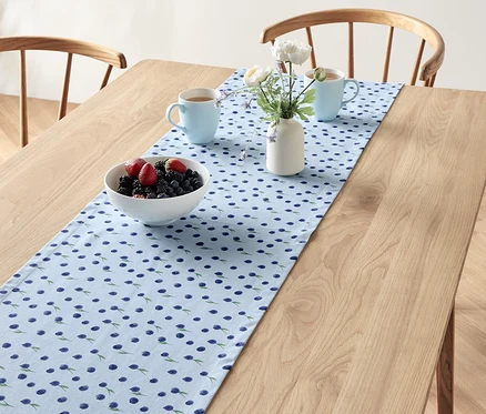 Auf einem Holztisch liegt ein Tischläufer mit Blaubeermuster, darauf eine Schale mit Beeren, zwei blaue Tassen und eine Vase mit Blumen.