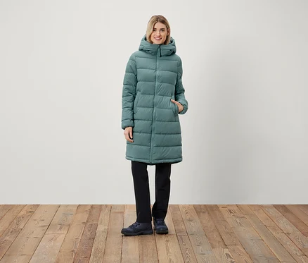Eine Frau steht und trägt einen langen Steppmantel mit Kapuze, eine schwarze Hose und Schuhe.