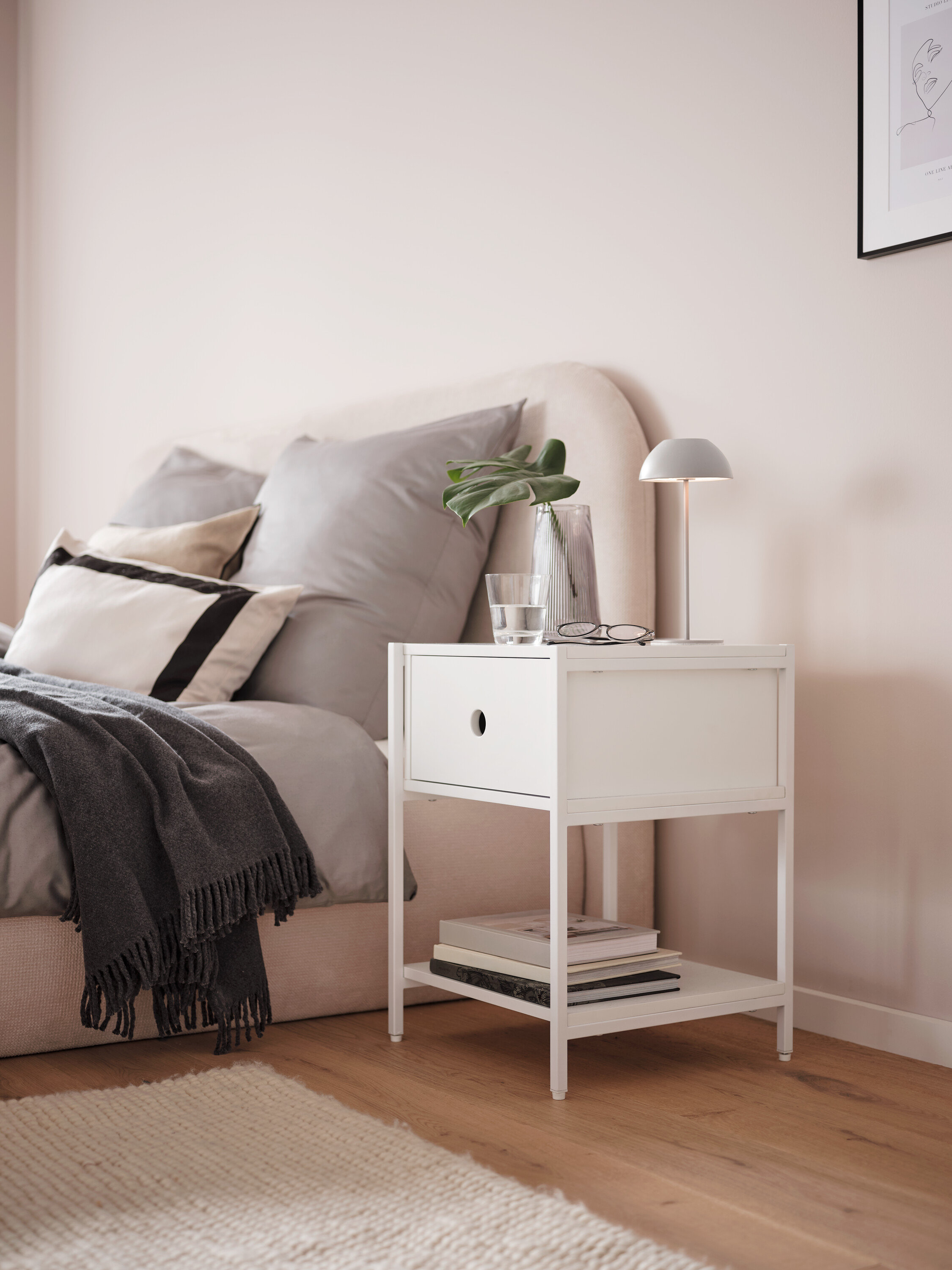 Eine graue LED-Akku-Tischleuchte steht auf einem weißen Nachttisch neben einem Bett.