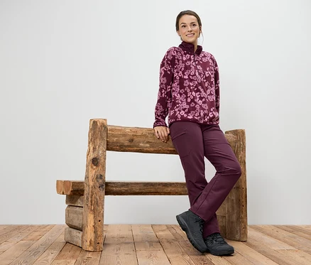 Eine Frau in einem purpurroten Fleecepullover mit Blumenmuster und Hose lehnt an einer Holzbank.