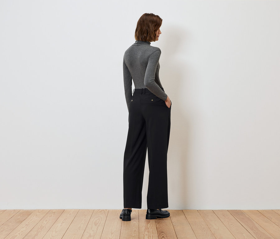 Rückansicht einer Frau mit den Händen in den Taschen einer schwarzen Hose und einem grauen Rollkragen.