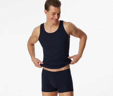 Lächelnder Mann in einem blauen SCHIESSER Feinripp-Unterhemd und blauen Boxershorts, der das Unterhemd mit beiden Händen hochzieht.
