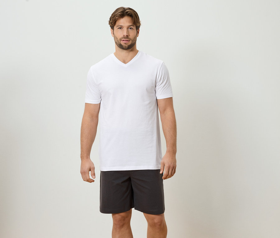 Mann steht vor einer weißen Wand und trägt ein weißes T-Shirt und graue Shorts.