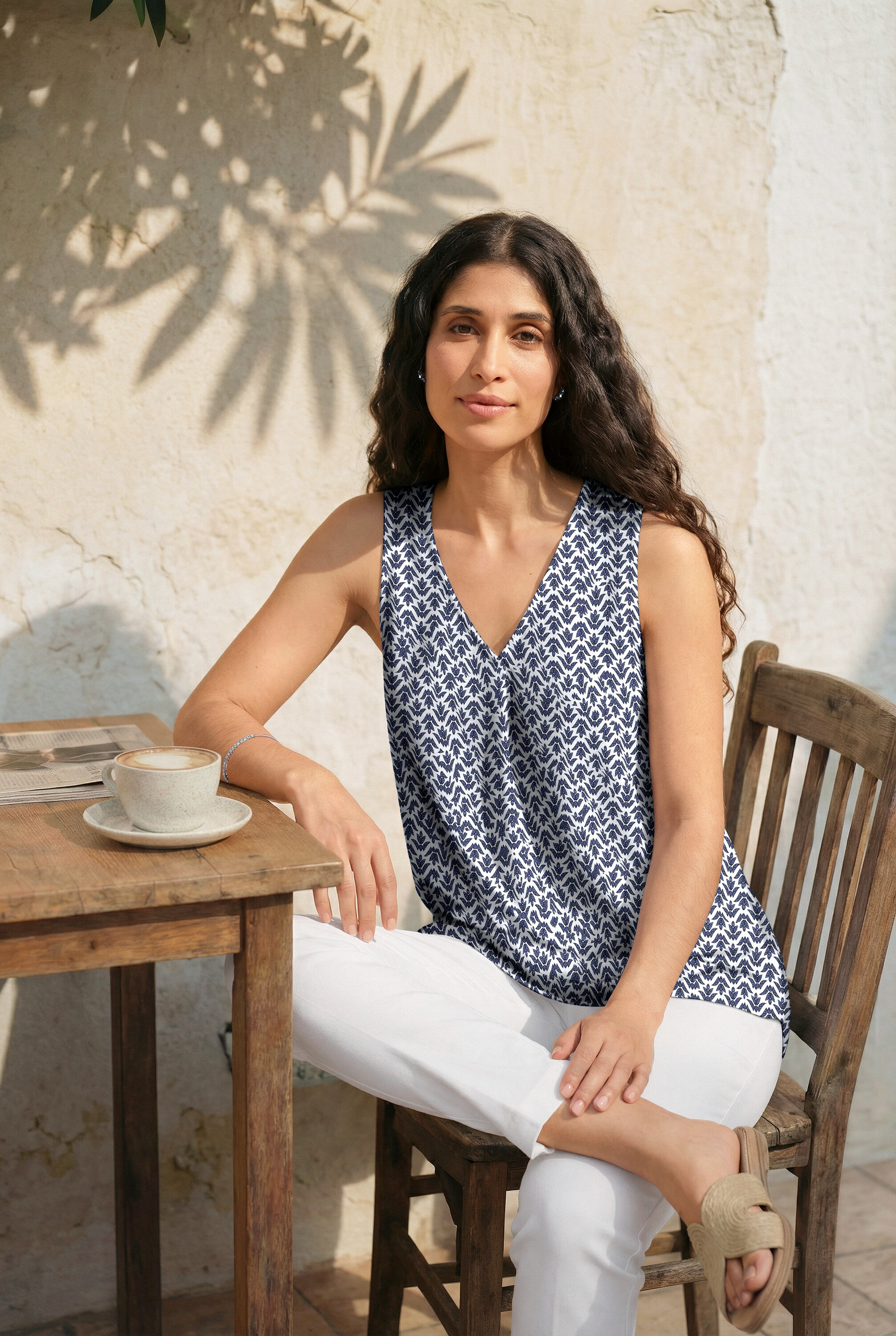 Frau sitzt am Tisch mit Ohrsteckern, Tennisarmband. Sie trägt ein Top mit Minimalprint, 7/8-Stretchhose und Pantoletten.