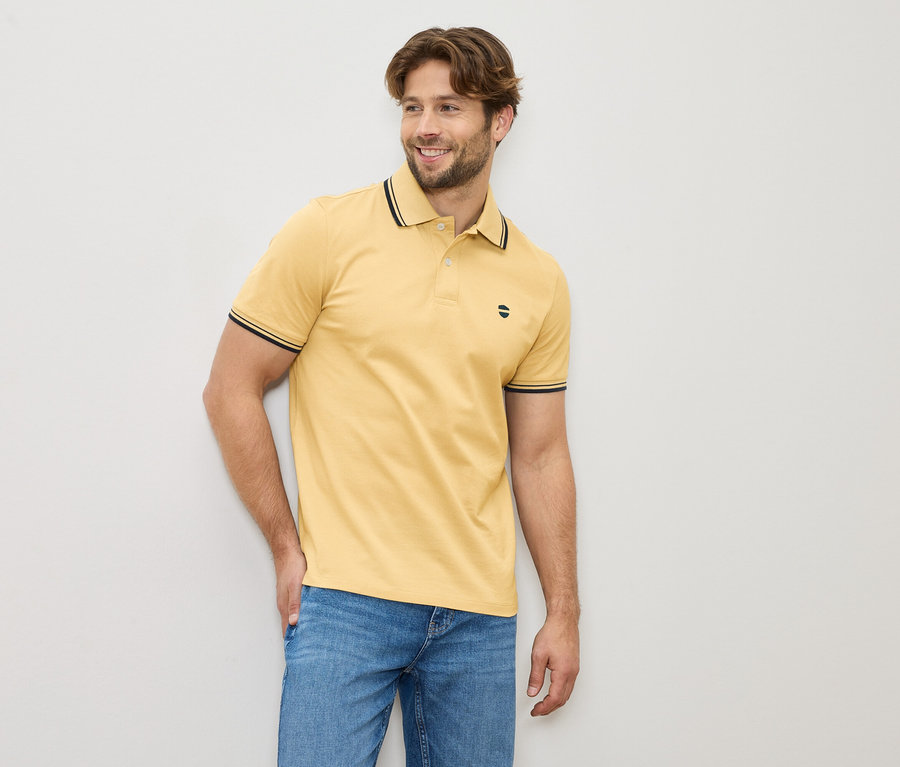 Mann mit gelbem Poloshirt und Jeans, stehend vor einer hellen Wand.