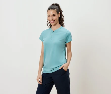 Frau mit lockigem Haar und Pferdeschwanz, die ein türkisfarbenes Shirt und marineblaue Hosen trägt.