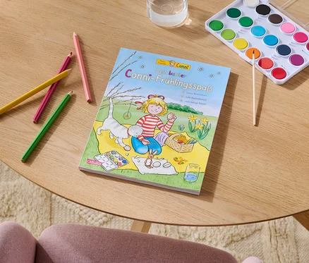 Auf einem Tisch liegen Buntstifte, ein Malbuch namens "Conni-Frühlingsspaß", ein Wasserfarbkasten mit Pinsel und ein Glas Wasser.