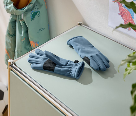 Auf einem Tisch liegen zwei blaue Handschuhe mit schwarzen Handflächen. Im Hintergrund sind eine Jacke mit Dinosaurier-Muster und ein Fußball zu sehen.