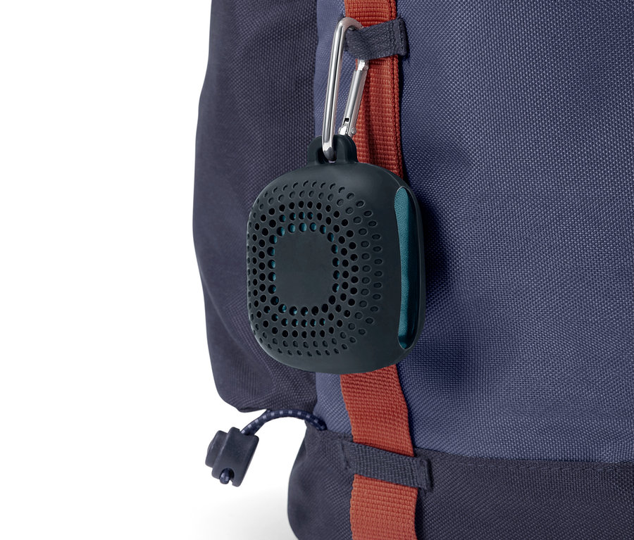 Eine schwarze, runde Hülle mit Löchern hängt mit einem Karabiner an einem blauen Rucksack.