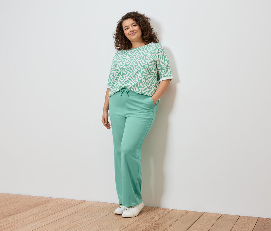 Frau posiert an einer Wand, trägt ein hellgrün gemustertes Oberteil, hellgrüne Hosen und weiße Turnschuhe.