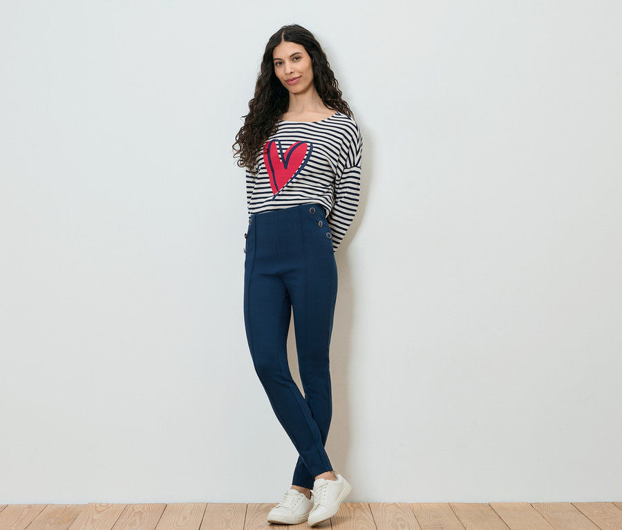 Frau mit dunklen, lockigen Haaren, gekleidet in ein gestreiftes Langarmshirt mit einem roten Herz darauf, marineblaue Hose und weiße Turnschuhe, posiert vor einer weißen Wand.