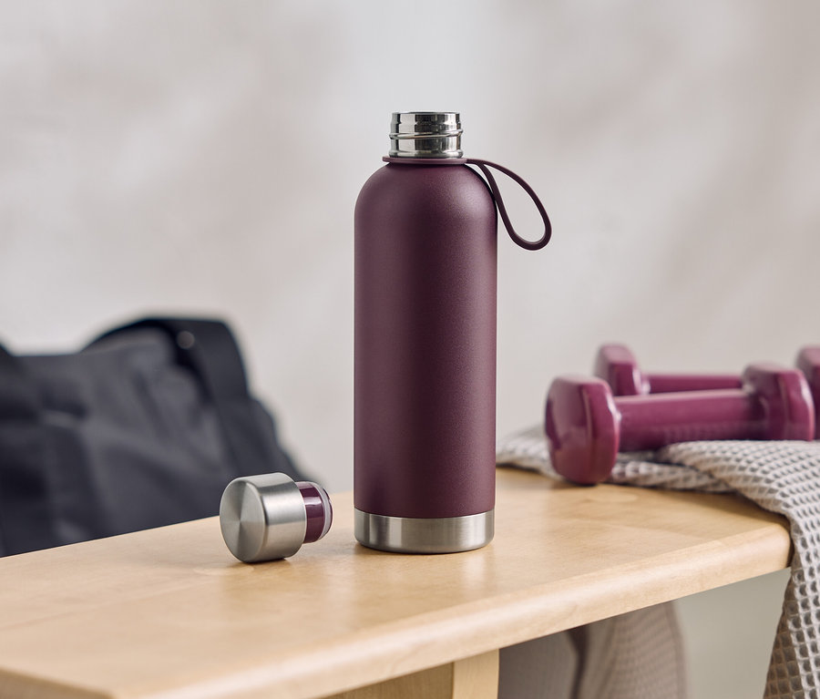 Eine auberginefarbene Wasserflasche steht mit geöffnetem Deckel neben Hanteln und einer Sporttasche auf einer Holzbank.