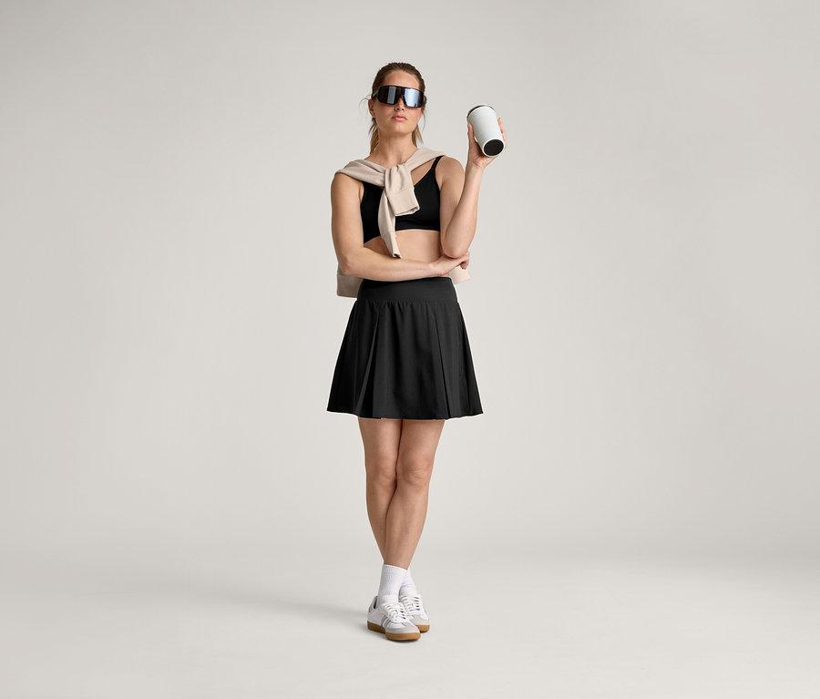 Frau mit Sonnenbrille, Sport-BH, Rock, Socken und Turnschuhen hält einen Becher in der Hand, während ein Pullover über ihre Schultern hängt.