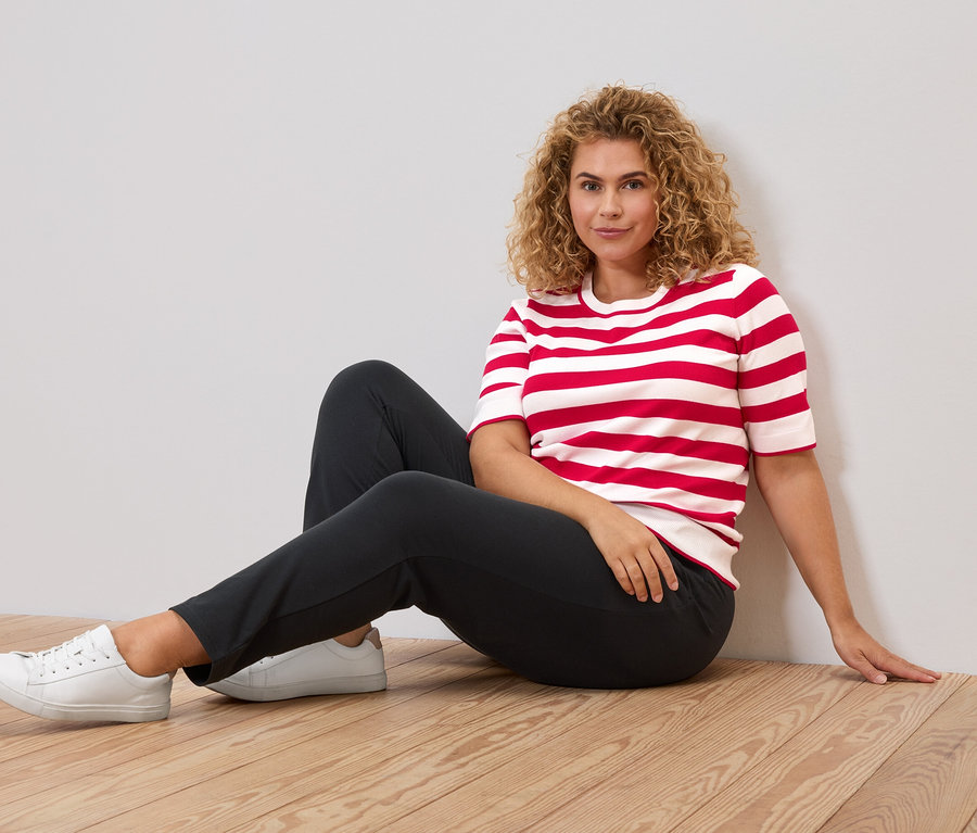 Frau mit lockigen Haaren sitzt auf einem Holzboden, trägt ein rot-weiß gestreiftes T-Shirt, dunkle Hose und weiße Turnschuhe.