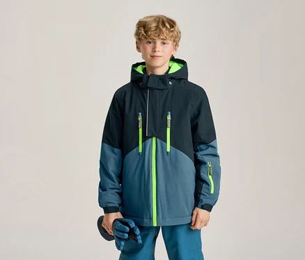 Junger Junge steht, trägt eine dunkelblaue und hellblaue Winterjacke mit neongrünen Reißverschlüssen und Kapuzenfutter, hält blaue Handschuhe.
