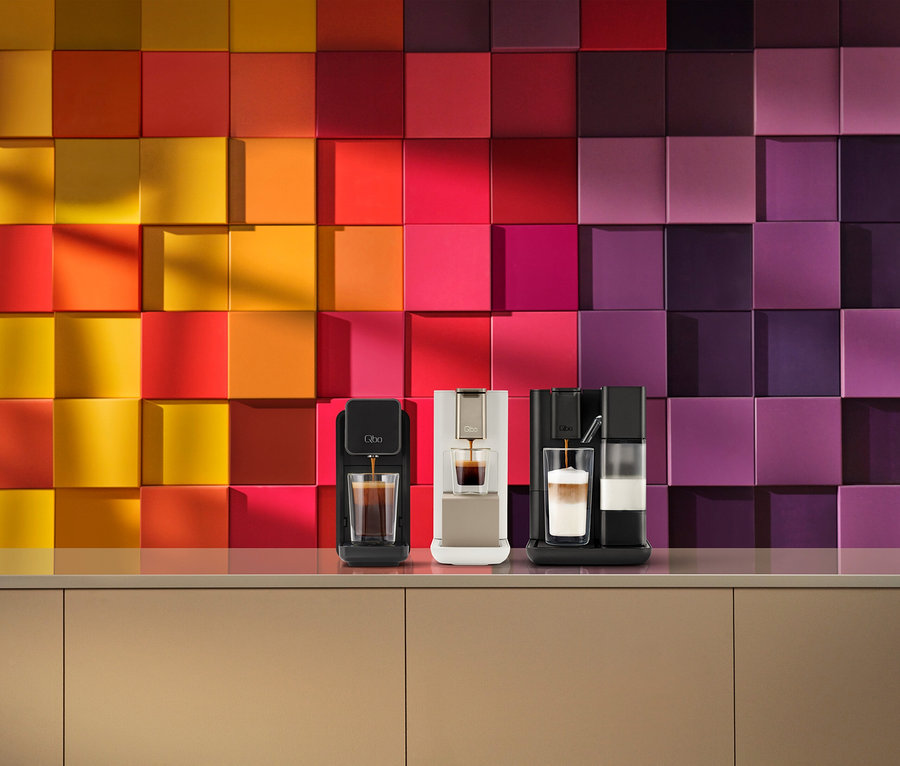 Drei Qbo Kaffeemaschinen auf einer Theke vor einer bunten Fliesenwand. Eine schwarze Qbo ONE Kapselmaschine brüht Kaffee, eine hellgoldene Qbo ICONIC brüht Kaffee und eine schwarze Qbo MILK MASTER mit Milchaufschäumer bereitet ein geschichtetes Getränk zu.