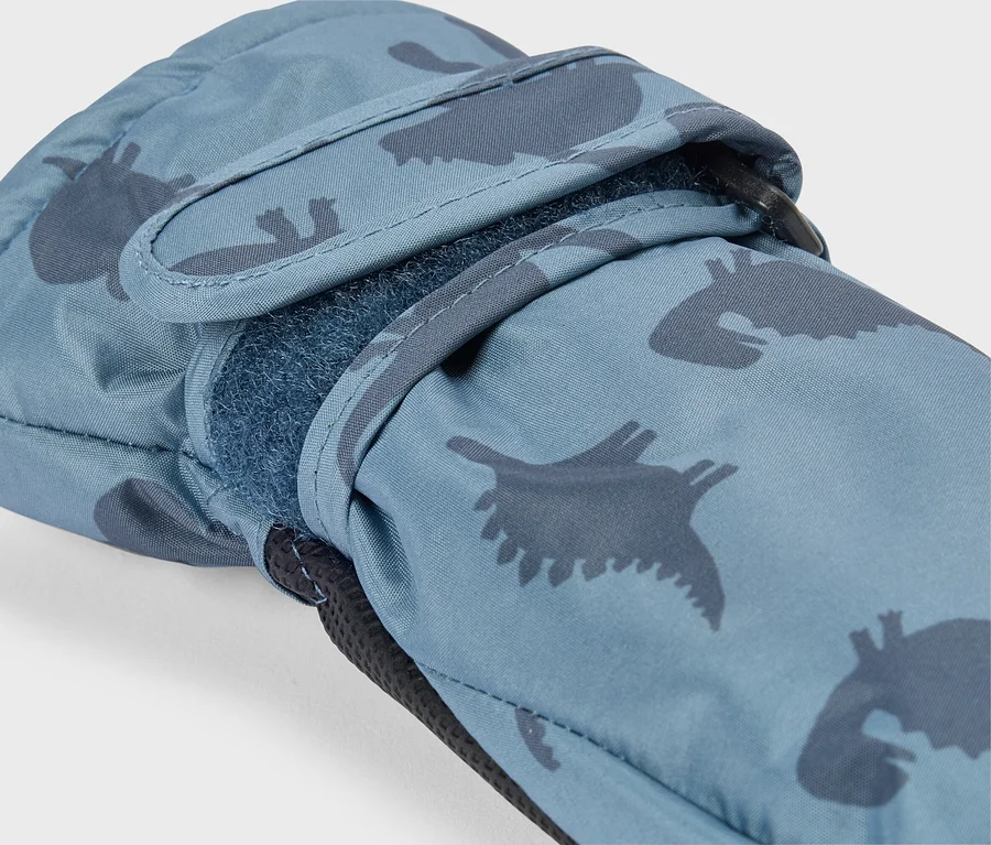 Nahaufnahme eines blauen Fäustlings mit dunklerem blauem Dinosaurier-Muster. Ein Klettverschluss am Handgelenk ist teilweise geöffnet und zeigt die flauschige Unterseite. Eine schwarze Schlaufe ist am Handgelenk sichtbar.
