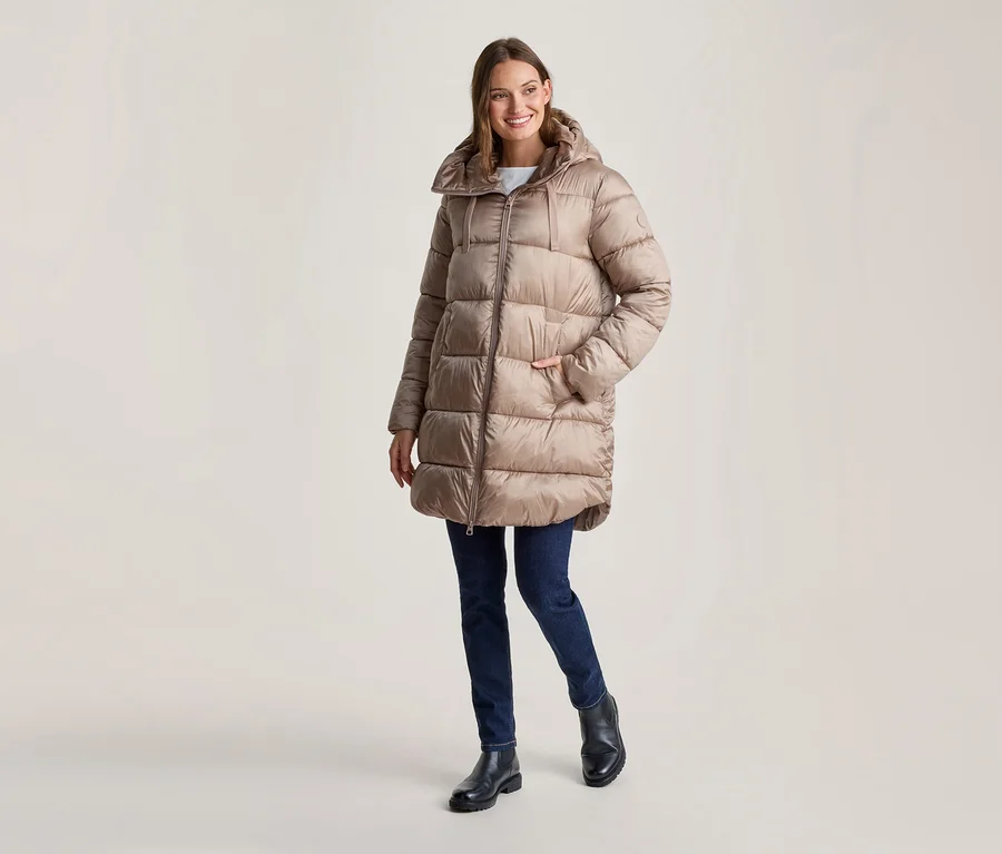 Lächelnde Frau in einem langen, beigen Kapuzen-Steppmantel, dunklen Jeans und schwarzen Stiefeln, eine Hand in der Tasche.