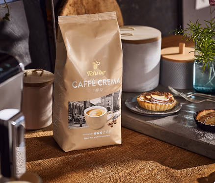 Auf einem Holztisch stehen eine Packung Caffè Crema Mild, eine Kaffeemaschine und Teller mit Kuchen.