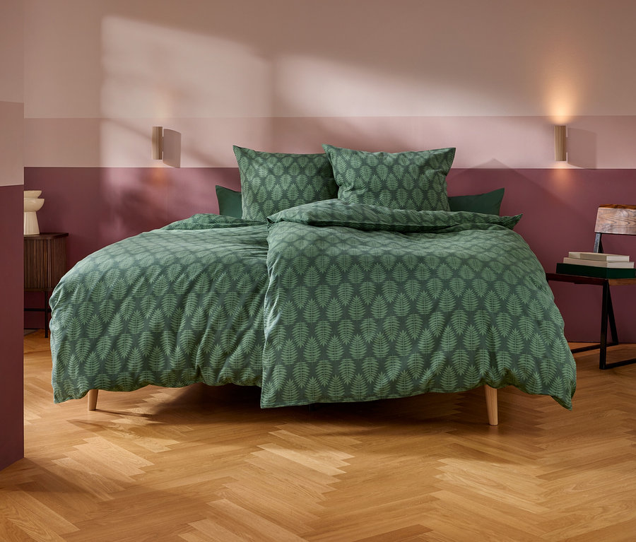 Ein Schlafzimmer mit einem Bett, das mit grüner Bettwäsche mit Blattmuster bezogen ist. Die Wände sind zweifarbig gestrichen, hellrosa oben und dunkelrotbraun unten. Zwei Wandleuchten erhellen den Raum. Links steht ein kleiner Holznachttisch mit einem weißen Dekoobjekt, rechts ein Holzstuhl mit Büchern. Der Boden ist aus hellem Holz im Fischgrätmuster.